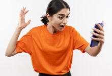 young girl wearing orange t shirt looking at her smartphone screen excited and confused with raised hand standing over white background