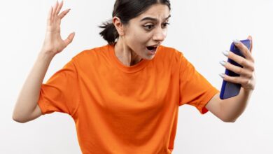 young girl wearing orange t shirt looking at her smartphone screen excited and confused with raised hand standing over white background