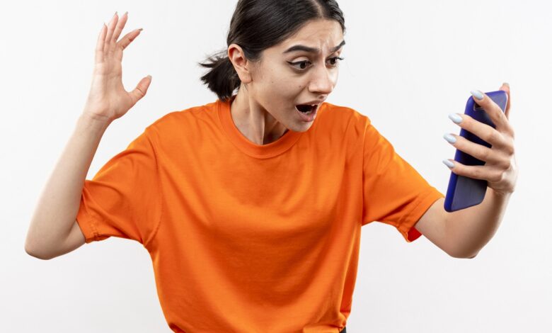 young girl wearing orange t shirt looking at her smartphone screen excited and confused with raised hand standing over white background