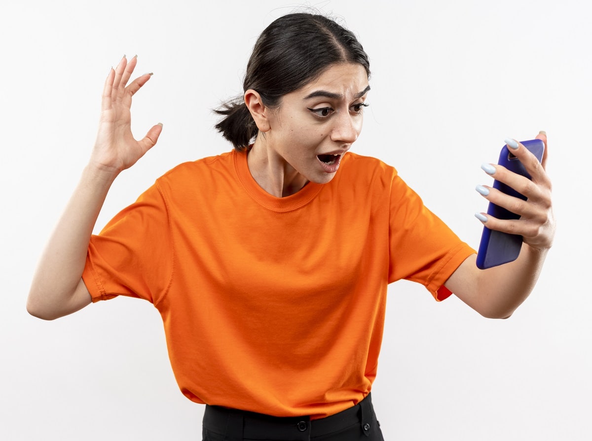 young girl wearing orange t shirt looking at her smartphone screen excited and confused with raised hand standing over white background