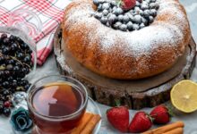 Bolo caseiro decorado com açúcar de confeiteiro e frutas vermelhas no topo, acompanhado de uma xícara de chá, paus de canela, morangos e groselhas, sobre uma tábua rústica de madeira.