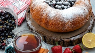 Bolo caseiro decorado com açúcar de confeiteiro e frutas vermelhas no topo, acompanhado de uma xícara de chá, paus de canela, morangos e groselhas, sobre uma tábua rústica de madeira.