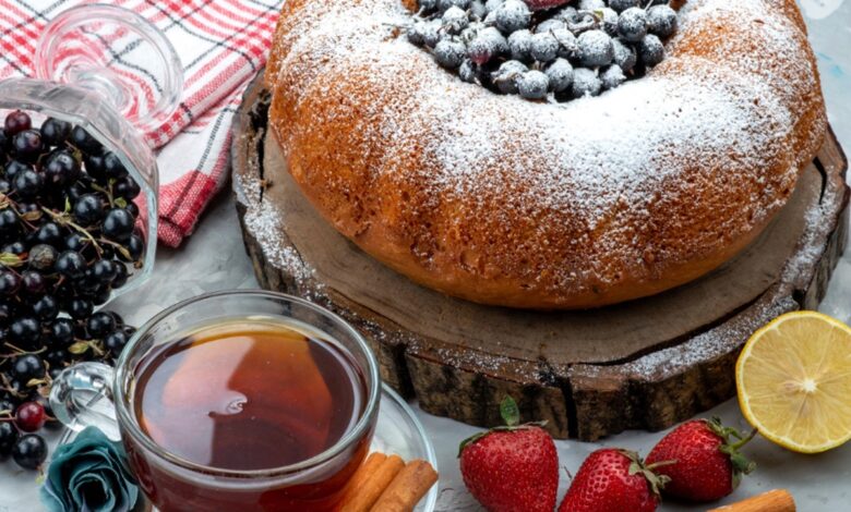 Bolo caseiro decorado com açúcar de confeiteiro e frutas vermelhas no topo, acompanhado de uma xícara de chá, paus de canela, morangos e groselhas, sobre uma tábua rústica de madeira.