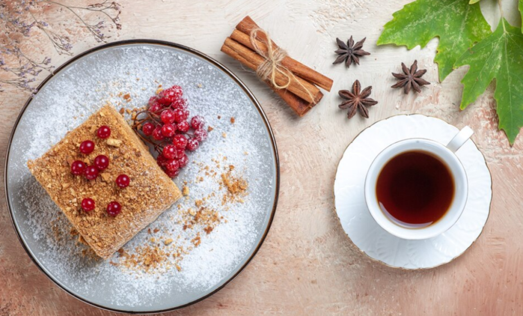 Prato com uma fatia de bolo coberta com açúcar de confeiteiro e decorada com frutas vermelhas, acompanhado de uma xícara de chá preto em um pires branco. Ao redor, paus de canela amarrados, anis-estrelado e folhas verdes sobre uma superfície rústica.