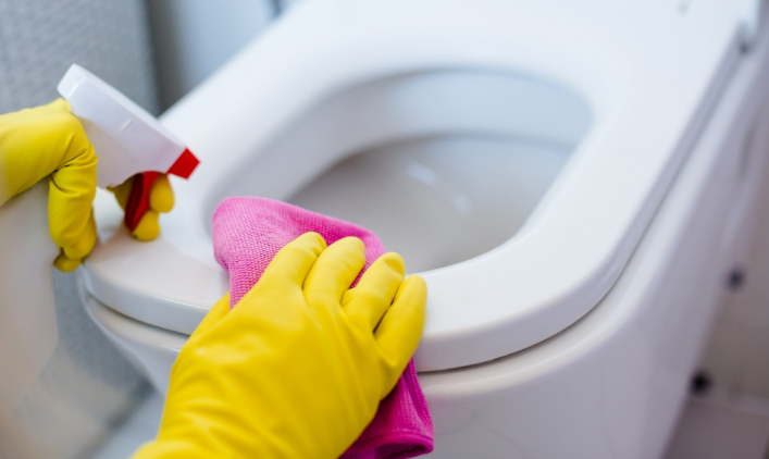 Pessoa utilizando um pano rosa para limpar a borda do vaso sanitário com luvas amarelas, seguindo truque de limpeza para manter o banheiro perfumado.