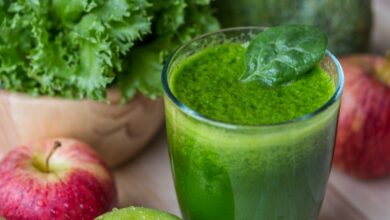 Suco verde de couve com maçã em um copo, com maçãs e folhas de couve ao fundo.