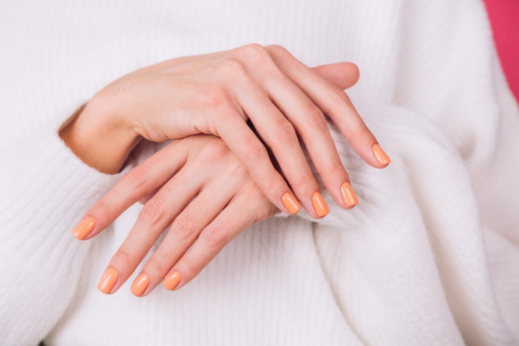 Mãos femininas com unhas pintadas em tom de pêssego brilhante, sobre um suéter branco.