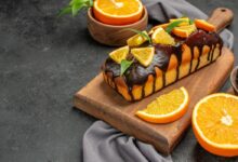 Bolo de laranja fofinho com calda de chocolate e rodelas de laranja sobre uma tábua de madeira.