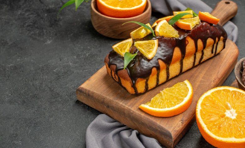 Bolo de laranja fofinho com calda de chocolate e rodelas de laranja sobre uma tábua de madeira.