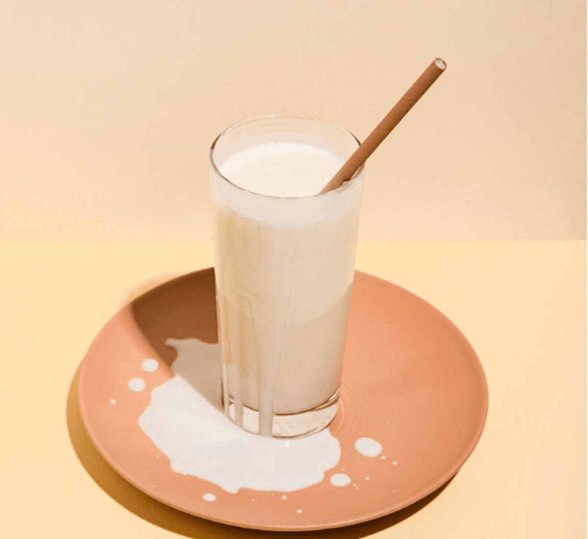 Copo de vitamina de mandioca servido com canudo, sobre uma base de prato com leite derramado