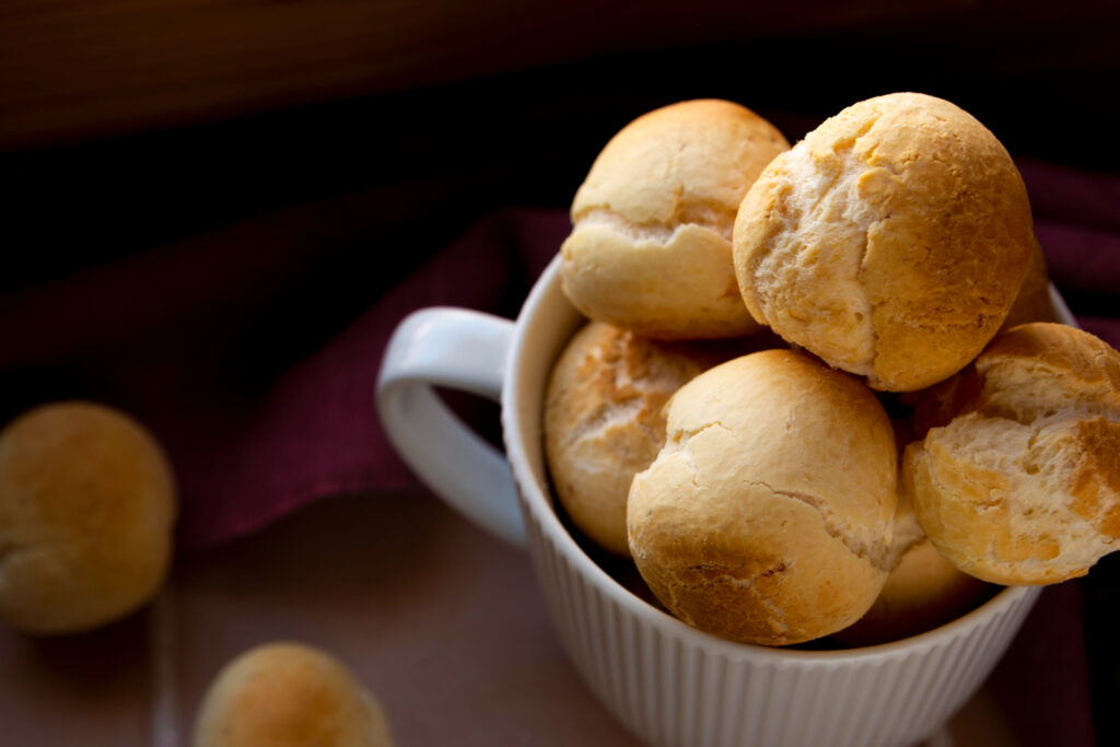  Pães de queijo dourados servidos em uma xícara branca, com fundo rústico desfocado