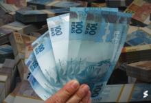 Mão segurando notas de 100 reais com pilhas de dinheiro ao fundo.