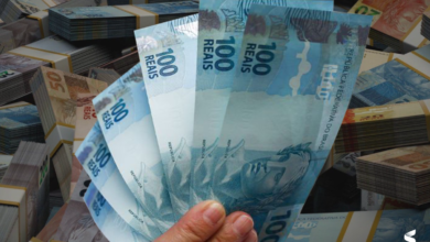 Mão segurando notas de 100 reais com pilhas de dinheiro ao fundo.