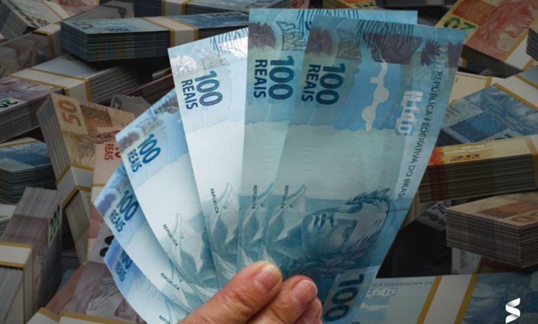 Mão segurando notas de 100 reais com pilhas de dinheiro ao fundo.