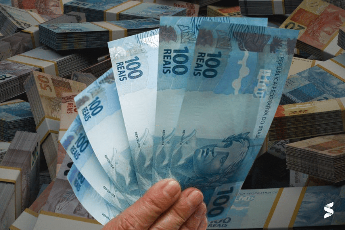 Mão segurando notas de 100 reais com pilhas de dinheiro ao fundo.