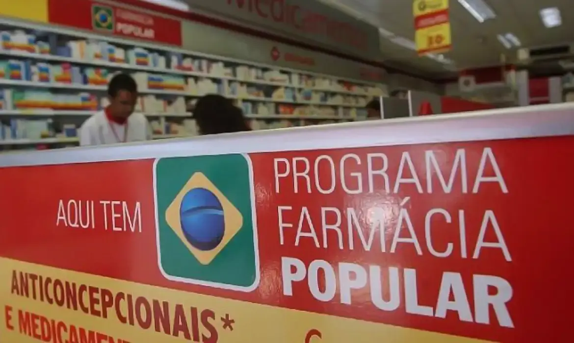 Placa do Programa Farmácia Popular em uma farmácia, com medicamentos visíveis ao fundo.