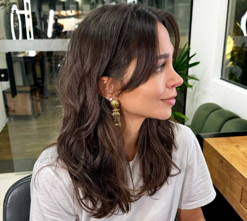 Mulher com cabelo castanho médio ondulado e franja lateral longa, destacando um corte sofisticado.