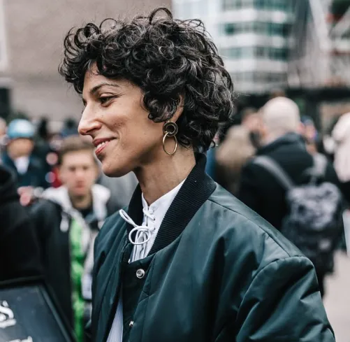 Mulher com cabelo curto e cacheado, usando um casaco, com um sorriso confiante em um ambiente urbano.