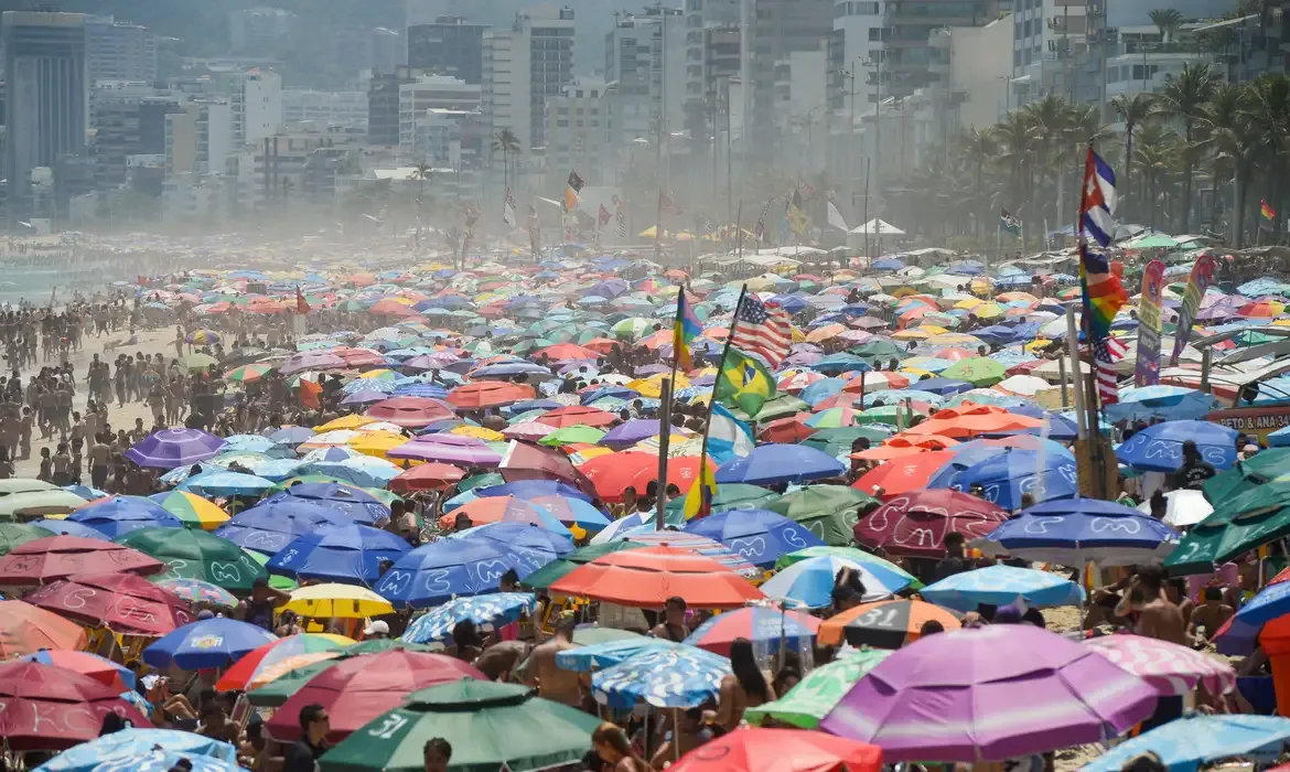 Multidão na praia do Rio de Janeiro, com várias sombrinhas coloridas, em um dia de calor extremo.