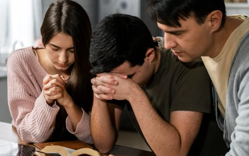 Três pessoas, em um momento de oração em família, com as mãos unidas em um livro sagrado.