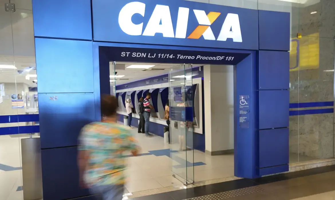 Entrada de uma agência da Caixa Econômica Federal com pessoas utilizando os caixas eletrônicos.