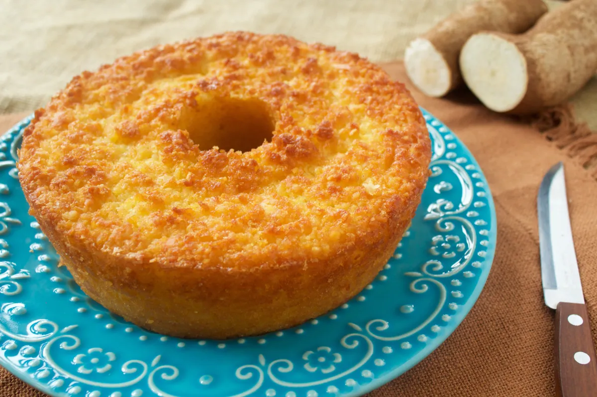 Bolo de mandioca redondo, com crosta dourada e uma textura macia, acompanhado por pedaços de mandioca.