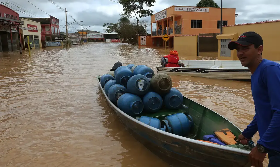 Homem em barco transportando botijões de gás em meio a uma enchente em área urbana.