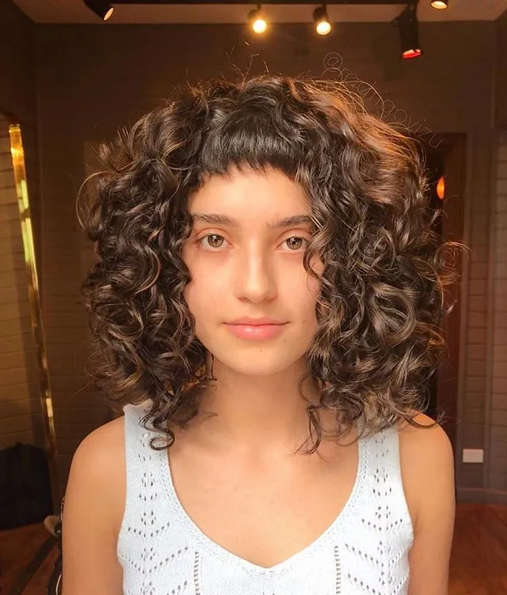 Mulher com cabelo cacheado e franja, usando um top branco, com um visual natural e moderno.