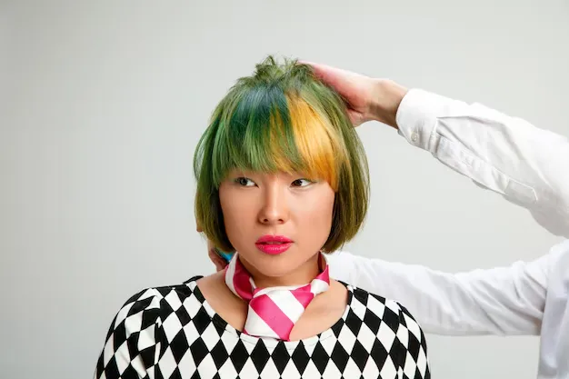 Mulher com cabelo colorido em verde e amarelo, usando uma blusa com padrão de losangos e lenço rosa.