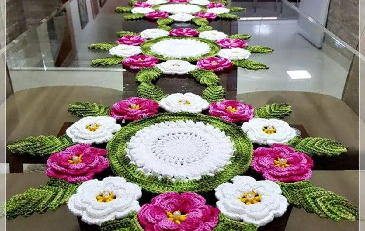 Caminho de mesa de crochê com flores coloridas em tons de rosa e branco, com detalhes verdes, em uma mesa.