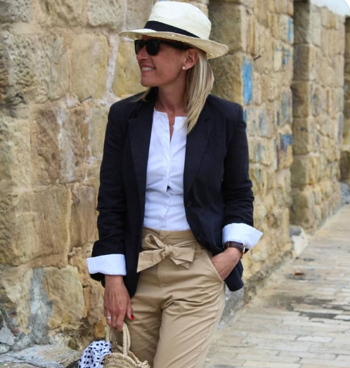 Mulher sorrindo usando chapéu de palha, blusa branca, blazer preto e calça bege, caminhando em uma rua com paredes de pedra.