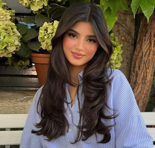 Mulher com cabelos longos, escuros e volumosos, usando uma camisa azul e sorrindo suavemente.