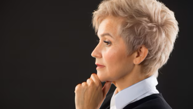 Mulher elegante com corte de cabelo pixie curto e loiro, vestindo blazer preto, em um fundo escuro.