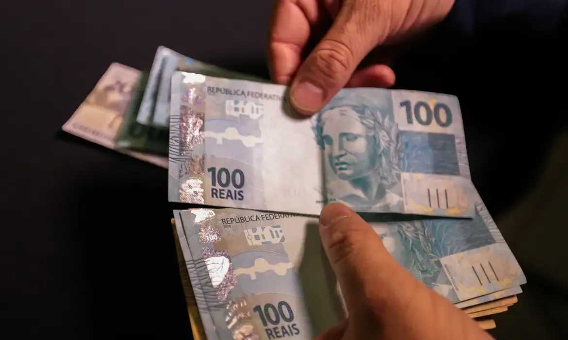 Mãos segurando notas de 100 reais, simbolizando investigações sobre a renda per capita de famílias.