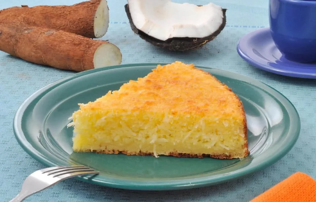 Fatia de bolo de mandioca dourado servida em um prato verde, acompanhada de mandioca e coco ao fundo