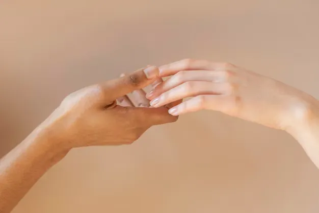 Duas mãos se tocando, uma com unhas naturais e a outra com esmalte nude, sobre fundo neutro.