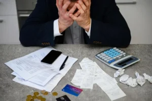 Homem preocupado com as finanças, lidando com recibos e documentos, simbolizando as dificuldades financeiras após o falecimento de um titular.