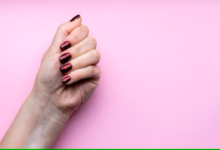 Mão com unhas bem feitas, pintadas com esmalte escuro e brilhante, sobre fundo rosa.