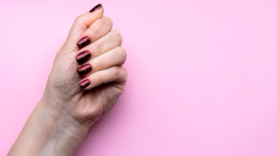 Mão com unhas bem feitas, pintadas com esmalte escuro e brilhante, sobre fundo rosa.