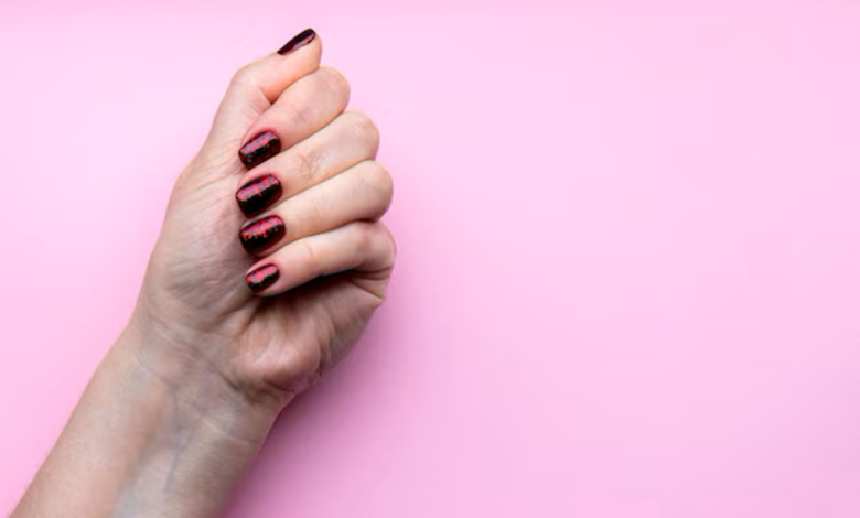 Mão com unhas bem feitas, pintadas com esmalte escuro e brilhante, sobre fundo rosa.