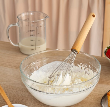 Creme batido em uma tigela de vidro com fouet de madeira, ao lado de um medidor com leite.