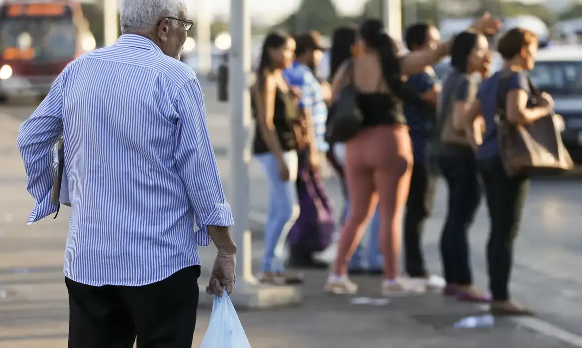 Idoso com sacola caminhando na rua, observando um grupo de pessoas.