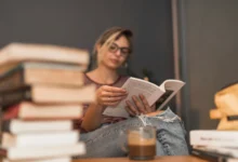 Mulher lendo um livro em um ambiente aconchegante, com pilhas de livros e uma xícara de café ao fundo