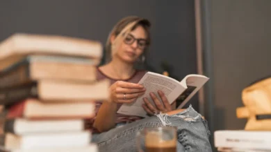 Mulher lendo um livro em um ambiente aconchegante, com pilhas de livros e uma xícara de café ao fundo