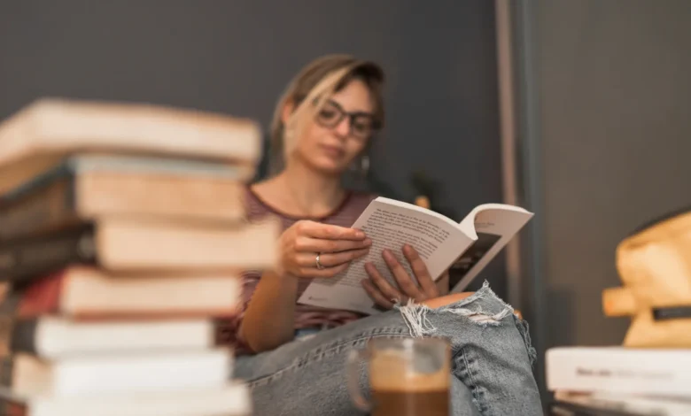 Mulher lendo um livro em um ambiente aconchegante, com pilhas de livros e uma xícara de café ao fundo