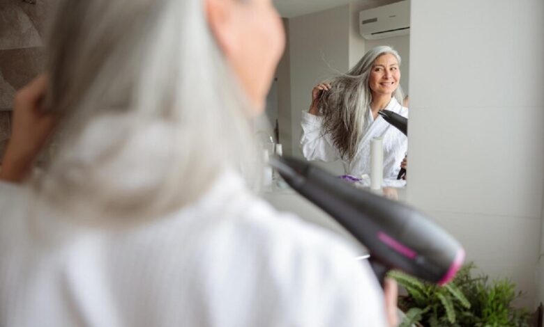 Mulher com cabelo grisalho secando os fios no espelho, representando os cuidados ao alisar cabelos grisalhos.