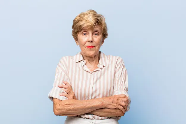 Mulher idosa com cabelo loiro curto, vestindo camisa listrada, com expressão confiante e braços cruzados.