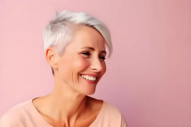 Mulher sorridente com cabelo pixie platinado, vestindo blusa rosa, contra um fundo rosa.