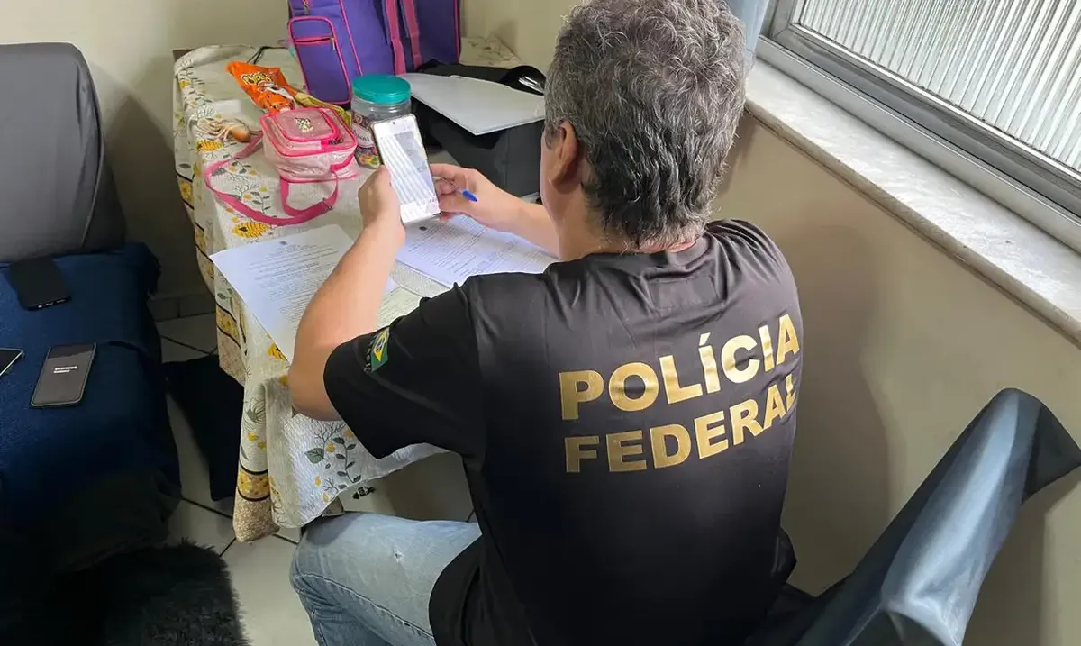 Agente da Polícia Federal analisando documentos e um celular durante operação.