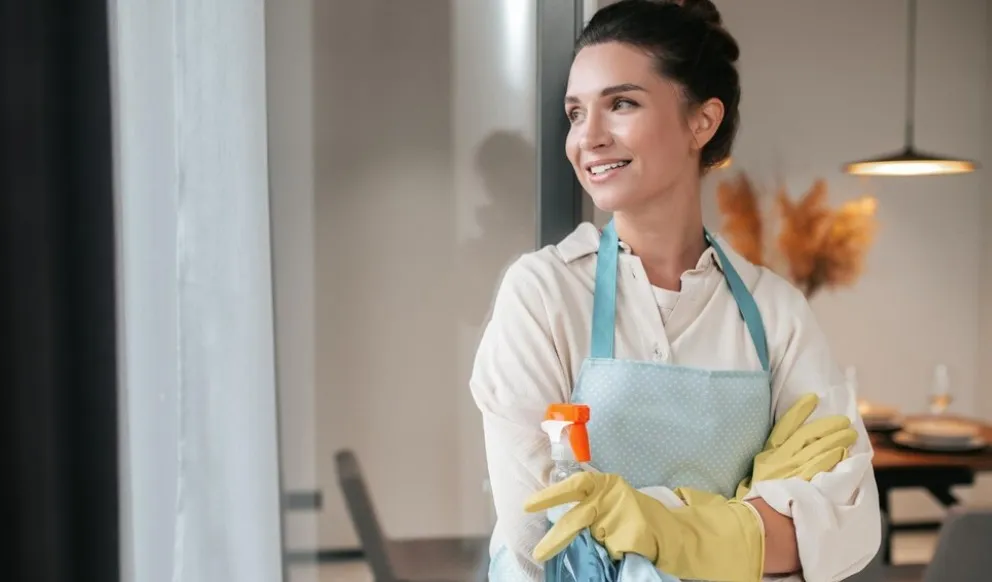 Mulher sorridente com luvas de limpeza e avental, segurando produtos de limpeza.